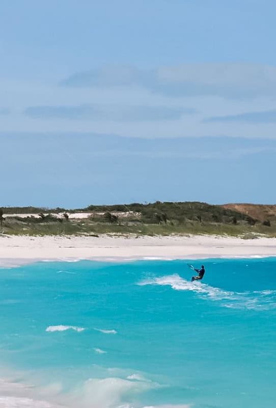 explore-abaco-2 | The Sandpiper Inn & Cottages, South Abaco, The Bahamas Kiteboarder riding a turquoise wave near a white-sand beach with low dunes under a blue sky.