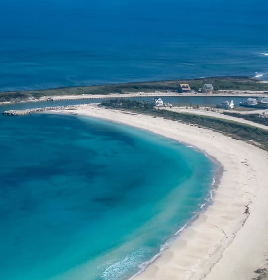 welcome-img | The Sandpiper Inn & Cottages, South Abaco, The Bahamas Aerial view of a crescent-shaped white-sand beach with vivid turquoise water and a small road with houses along the coast.